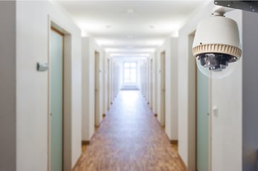 A long hallway in an apartment complex with a window at the end with several light green doors on either side. On the right side of the hall is a closeup view of a security camera in a glass case.