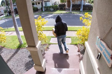 A view from a doorbell security camera of the front porch of a home with a person in a black hoodie and jeans walking away from the porch with the homeowner's package. 
